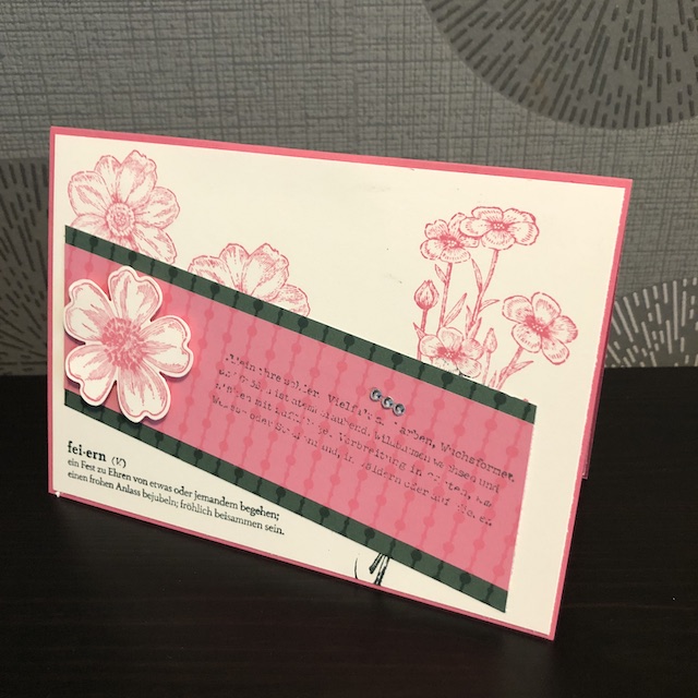 Zu sehen ist eine Pop Up Karte mit Flügeln. Auf der rosanen Grundkarte ist ein Auflieger in Vanille Pur geklebt. Dadrauf sind verschiedene Blüten in rosa gestempelt. Quer darüber liegen zwei Papierstreifen in dunkelgrün und rosa auf denen eine erhabene Blüte geklebt ist. Unter den Streifen ist ein Spruch gestempelt, der lautet: fei-ern (v) ein Fest zu Ehren von etwas oder jemandem begehen; einen frohen Anlass bejubeln; fröhlich beisammen sein.