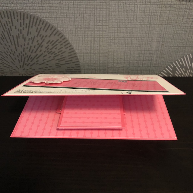 Zu sehen ist eine Pop Up Karte mit Flügeln. Auf der rosanen Grundkarte ist ein Auflieger in Vanille Pur geklebt. Dadrauf sind verschiedene Blüten in rosa gestempelt. Quer darüber liegen zwei Papierstreifen in dunkelgrün und rosa auf denen eine erhabene Blüte geklebt ist. Unter den Streifen ist ein Spruch gestempelt, der lautet: fei-ern (v) ein Fest zu Ehren von etwas oder jemandem begehen; einen frohen Anlass bejubeln; fröhlich beisammen sein.