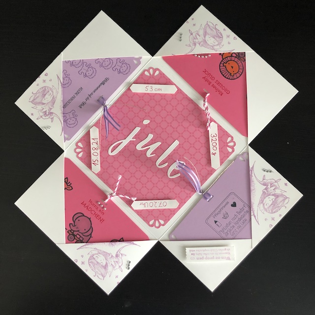 Die geöffnete Explosionsbox zur Geburt von Jule. In den vier Taschen stecken Karten mit Tieren und Grüßen bestempelt. In der Mitte sind die Buchstaben für den Namen Jule aufgeklebt. In allen Vier Ecken sind Schilder mit Gewicht, Länge, Uhrzeit und Geburtstag angebracht. Farblich finden wir hier Rosatöne, Flieder und Creme.