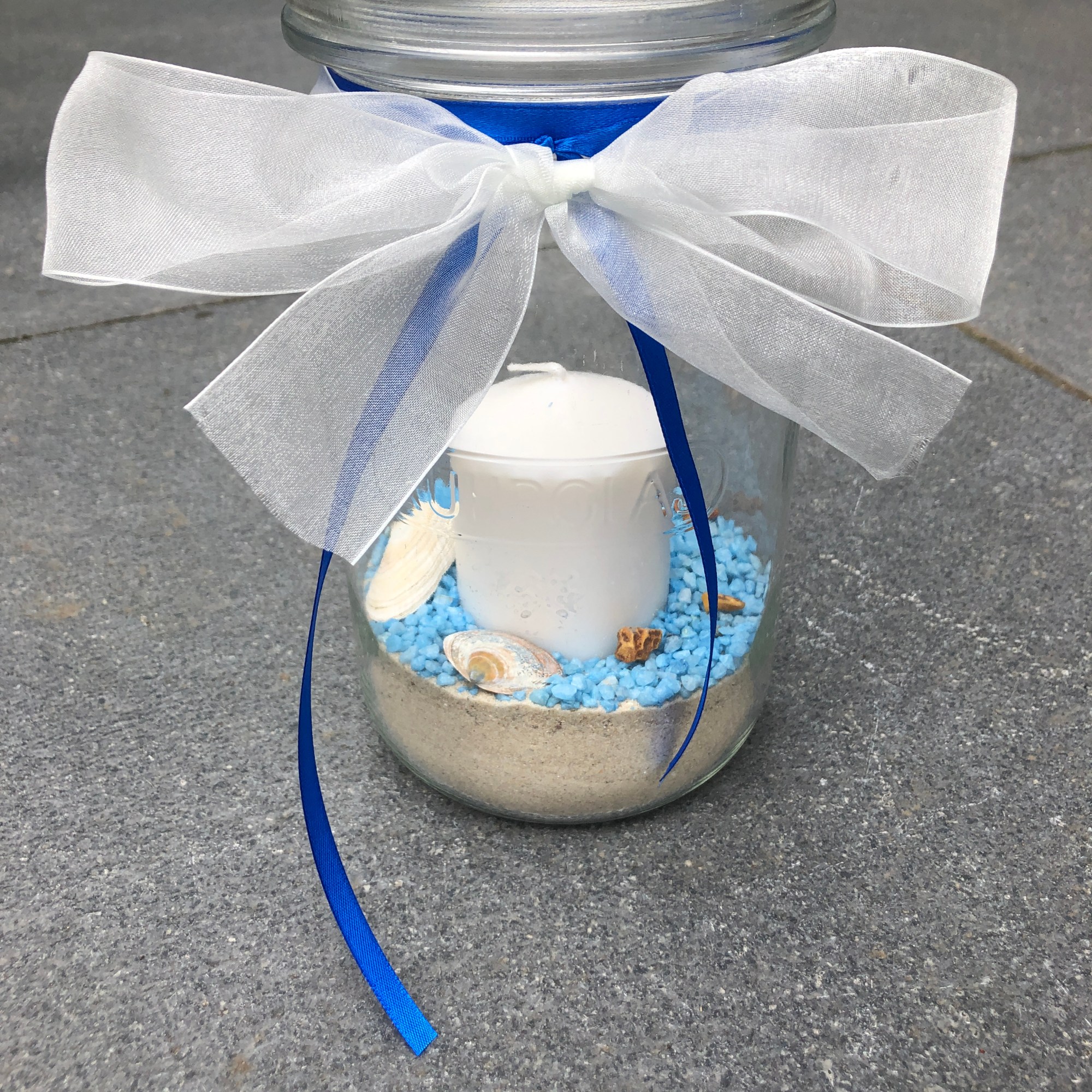 Ein 1,5l Einmachglas gefüllt mit Vogelsand. Darüber sind blaue Steinchen gestreut auf denen Muscheln und Schnecken aus dem Meer liegen. In der Mitte steckt eine weiße Stumpen Kerze. Das Weckglas ist oben mit einer weißen Taftschleife versehen und blauen Bändern.