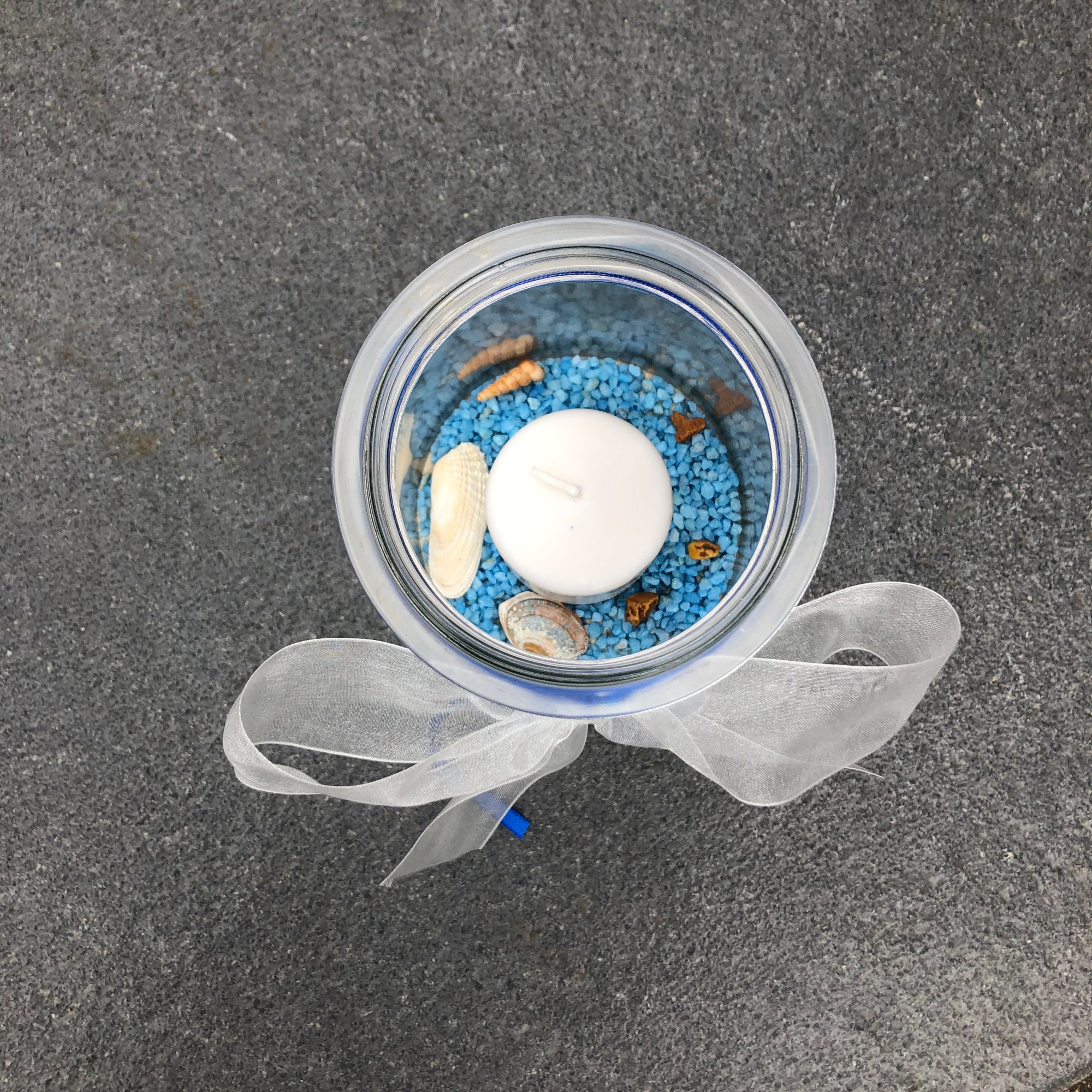 Sich von Oben: Ein 1,5l Einmachglas gefüllt mit Vogelsand. Darüber sind blaue Steinchen gestreut auf denen Muscheln und Schnecken aus dem Meer liegen. In der Mitte steckt eine weiße Stumpen Kerze. Das Weckglas ist oben mit einer weißen Taftschleife versehen und blauen Bändern.