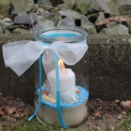 Ein 1,5l Einmachglas gefüllt mit Vogelsand. Darüber sind blaue Steinchen gestreut auf denen Muscheln und Schnecken aus dem Meer liegen. In der Mitte steckt eine weiße Stumpen Kerze. Das Weckglas ist oben mit einer weißen Taftschleife versehen und blauen Bändern.