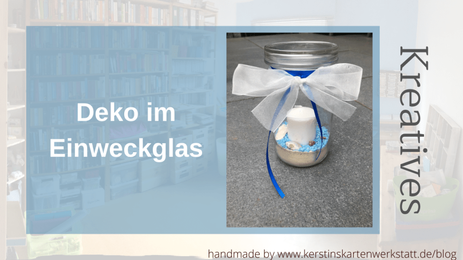 Ein 1,5l Einmachglas gefüllt mit Vogelsand. Darüber sind blaue Steinchen gestreut auf denen Muscheln und Schnecken aus dem Meer liegen. In der Mitte steckt eine weiße Stumpen Kerze. Das Weckglas ist oben mit einer weißen Taftschleife versehen und blauen Bändern.