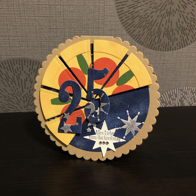 Runde Grußkarte zur Silbernen Hochzeit. Die Grundkarte ist bräunlich. Darauf bläuliches Papier mit Sternen. Zu zwei dritteln ist die Kreisförmige Karte als Pizza gestaltet auf der rote Schiben und grüne Stile angedeutet sind. Auf einem Stern steht der Gruß: Alles Liebe zum Hochzeitstag. Über der Pizza sind die Ziffern 25 angebracht.