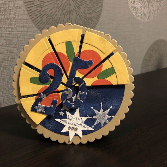 Runde Grußkarte zur Silbernen Hochzeit. Die Grundkarte ist bräunlich. Darauf bläuliches Papier mit Sternen. Zu zwei dritteln ist die Kreisförmige Karte als Pizza gestaltet auf der rote Schiben und grüne Stile angedeutet sind. Auf einem Stern steht der Gruß: Alles Liebe zum Hochzeitstag. Über der Pizza sind die Ziffern 25 angebracht.