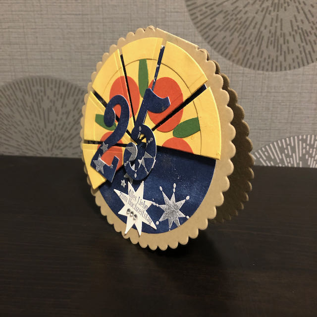 Runde Grußkarte zur Silbernen Hochzeit. Die Grundkarte ist bräunlich. Darauf bläuliches Papier mit Sternen. Zu zwei dritteln ist die Kreisförmige Karte als Pizza gestaltet auf der rote Schiben und grüne Stile angedeutet sind. Auf einem Stern steht der Gruß: Alles Liebe zum Hochzeitstag. Über der Pizza sind die Ziffern 25 angebracht.