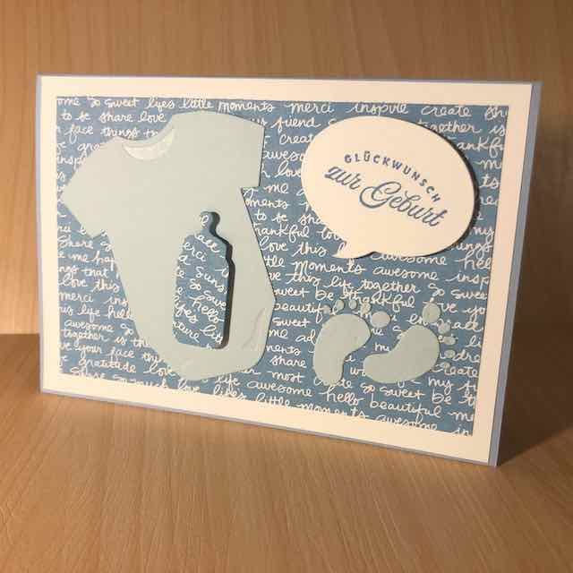 Pop Up Karte mit Flügeln zur Geburt eines Jungen in Blautönen. Auf der Karte ist ein hellblauer Babybody mit einer Trinkflasche in Designpapier zu sehen. Daneben ist ein Fußabdruck geklebt. Auf der Sprechblase ist der Spruch: Glückwunsch zur Geburt zu lesen.