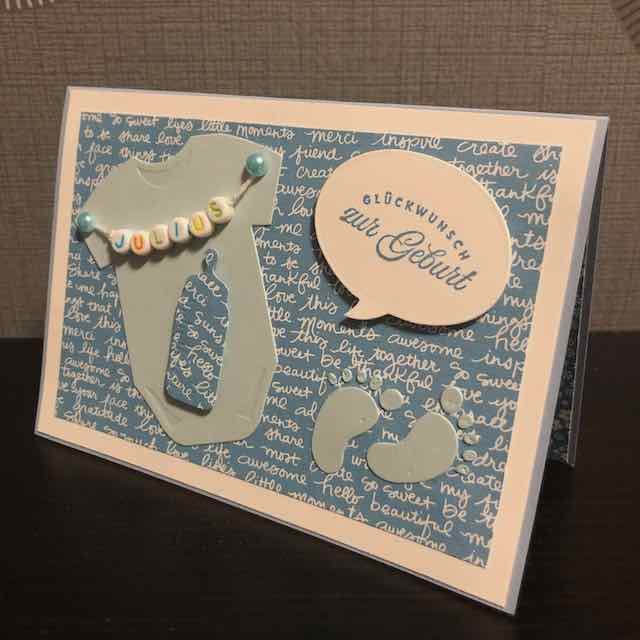 Pop Up Karte mit Flügeln zur Geburt eines Jungen in Blautönen. Auf der Karte ist ein hellblauer Babybody mit einer Trinkflasche in Designpapier zu sehen. Daneben ist ein Fußabdruck geklebt. Auf der Sprechblase ist der Spruch: Glückwunsch zur Geburt zu lesen.