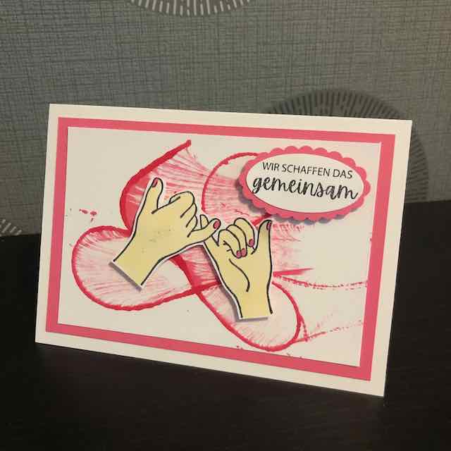 Grußkarte mit folgendem Text: Wir schaffen das gemeinsam. Zu sehen sind zwei Hände, die sich an den kleinen Fingern halten. Der Hintergrund wurde mit der Fadentechnik in rosa gestaltet.