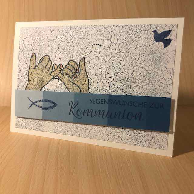 Grußkarte zur Kommunion, zwei Hände gestempelt, die sich an den kleinen Fingern festhalten, eine Friedenstaube in der oberen rechten Ecke, ein blauer Streifen mit Fisch und Spruch: Segenswünsche zur Kommunion