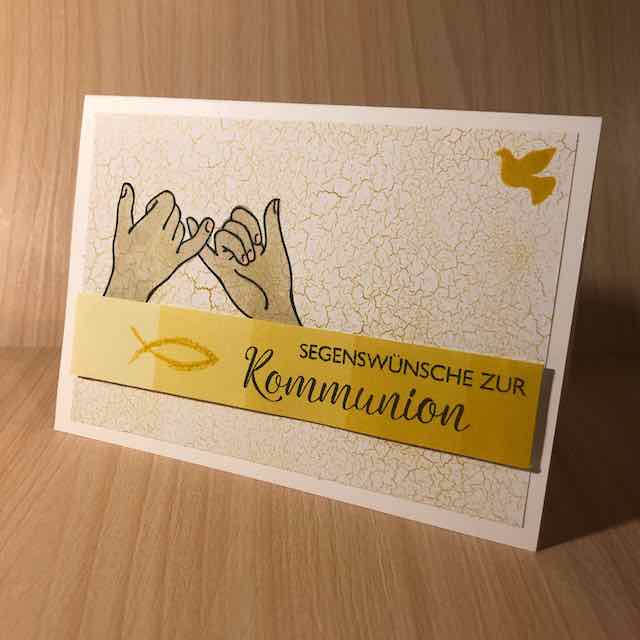 Grußkarte zur Kommunion, zwei Hände gestempelt, die sich an den kleinen Fingern festhalten, eine Friedenstaube in der oberen rechten Ecke, ein blauer Streifen mit Fisch und Spruch: Segenswünsche zur Kommunion