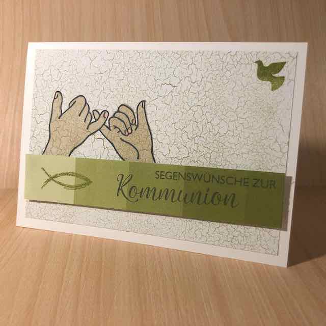 Grußkarte zur Kommunion, zwei Hände gestempelt, die sich an den kleinen Fingern festhalten, eine Friedenstaube in der oberen rechten Ecke, ein blauer Streifen mit Fisch und Spruch: Segenswünsche zur Kommunion