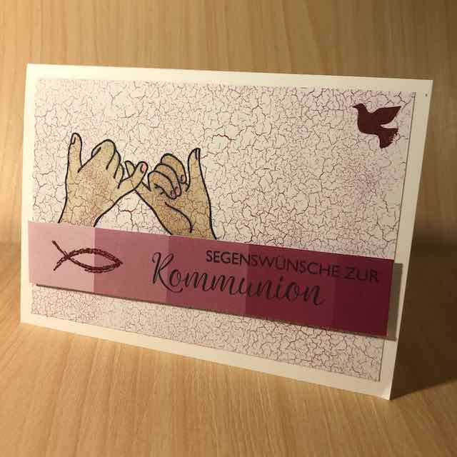 Grußkarte zur Kommunion, zwei Hände gestempelt, die sich an den kleinen Fingern festhalten, eine Friedenstaube in der oberen rechten Ecke, ein blauer Streifen mit Fisch und Spruch: Segenswünsche zur Kommunion
