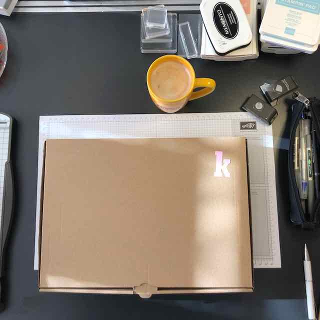 Foto von einem geschlossenen Karton in dem sich das Material für das Bastelwochenende befindet. Weiterhin sind Acrylblöcke, Stempelkissen, eine Federtasche und eine Tasse Kaffee zu sehen