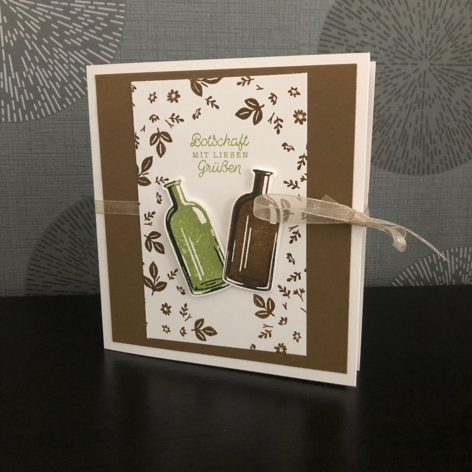 Eine kleine gebastelte Box im Buchformat. Vorne drauf sind zwei Weinflaschen aufgeklebt und der Spruch: Botschaft mit Lieben Grüßen. Die Box wird mit einem Band verschlossen.