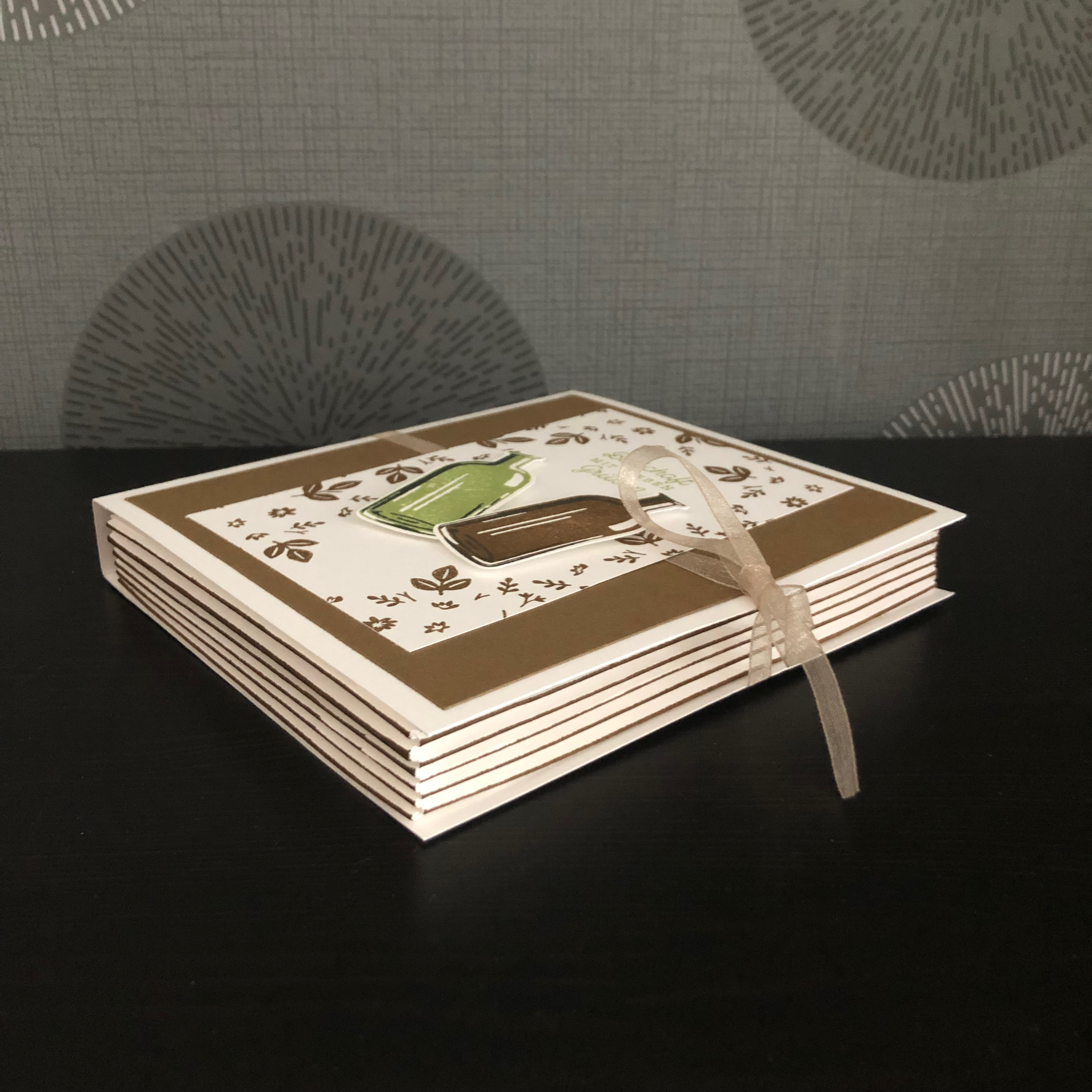 Eine kleine gebastelte Box im Buchformat. Vorne drauf sind zwei Weinflaschen aufgeklebt und der Spruch: Botschaft mit Lieben Grüßen. Die Box wird mit einem Band verschlossen.