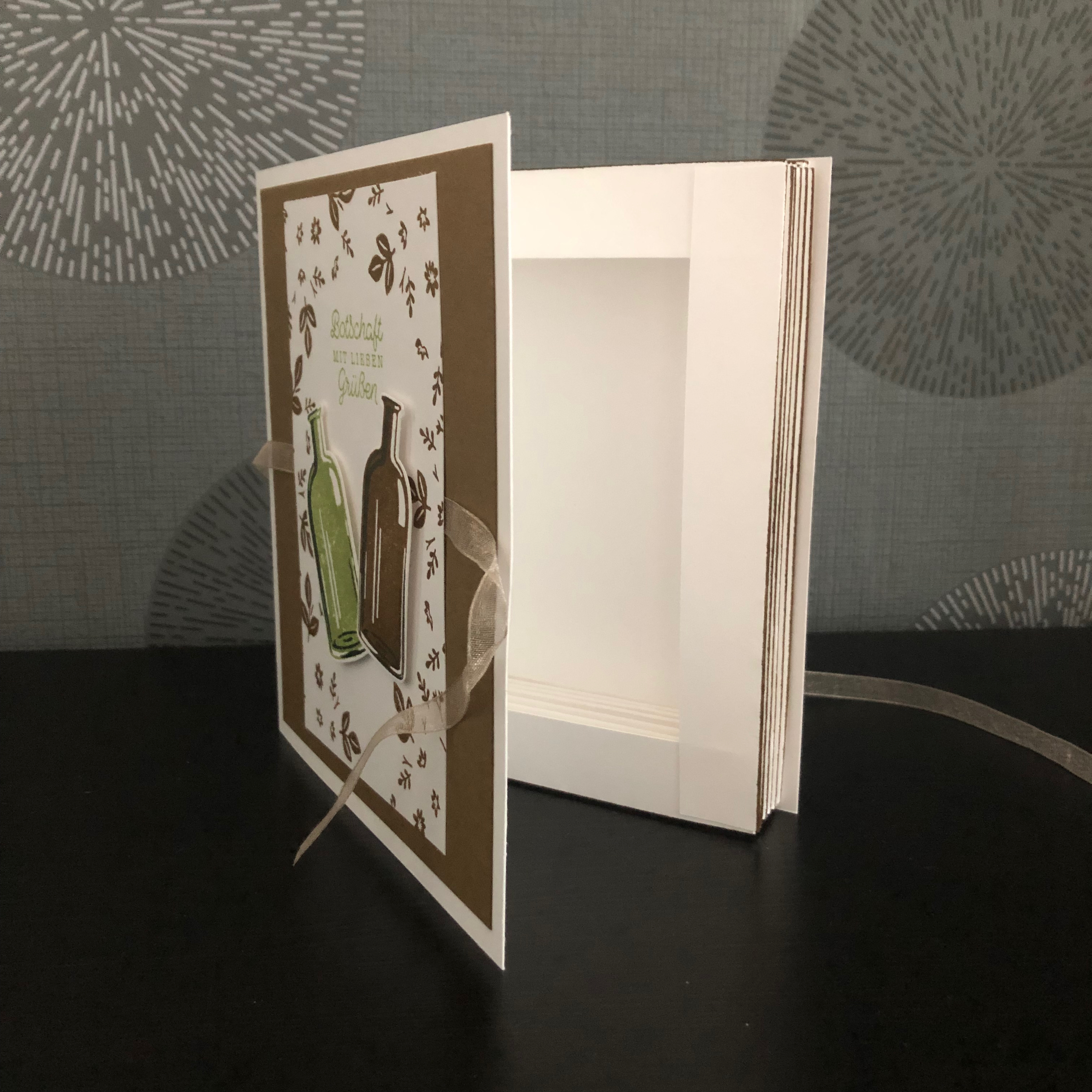Eine kleine gebastelte Box im Buchformat. Vorne drauf sind zwei Weinflaschen aufgeklebt und der Spruch: Botschaft mit Lieben Grüßen. Die Box wird mit einem Band verschlossen.
