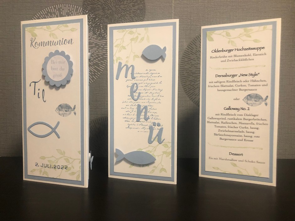 Zu sehen ist eine gebastelte Menükarte zur Kommunion in blau, weiß und grün für einen Jungen. Die Menükarte besteht aus drei Seiten. Die erste Seite ist beschriftet mit dem Namen des Kindes, das Datum der Kommunion und verziert mit zwei blauen Fischen. Die zweite Seite enthält das Wort Menü in ausgestanzten Buchstaben verzeiert mit zwei blauen Fischen und hinterlegt mit grünen Ranken und Schrift. Auf der dritten Seite ist das Menü gedruckt.