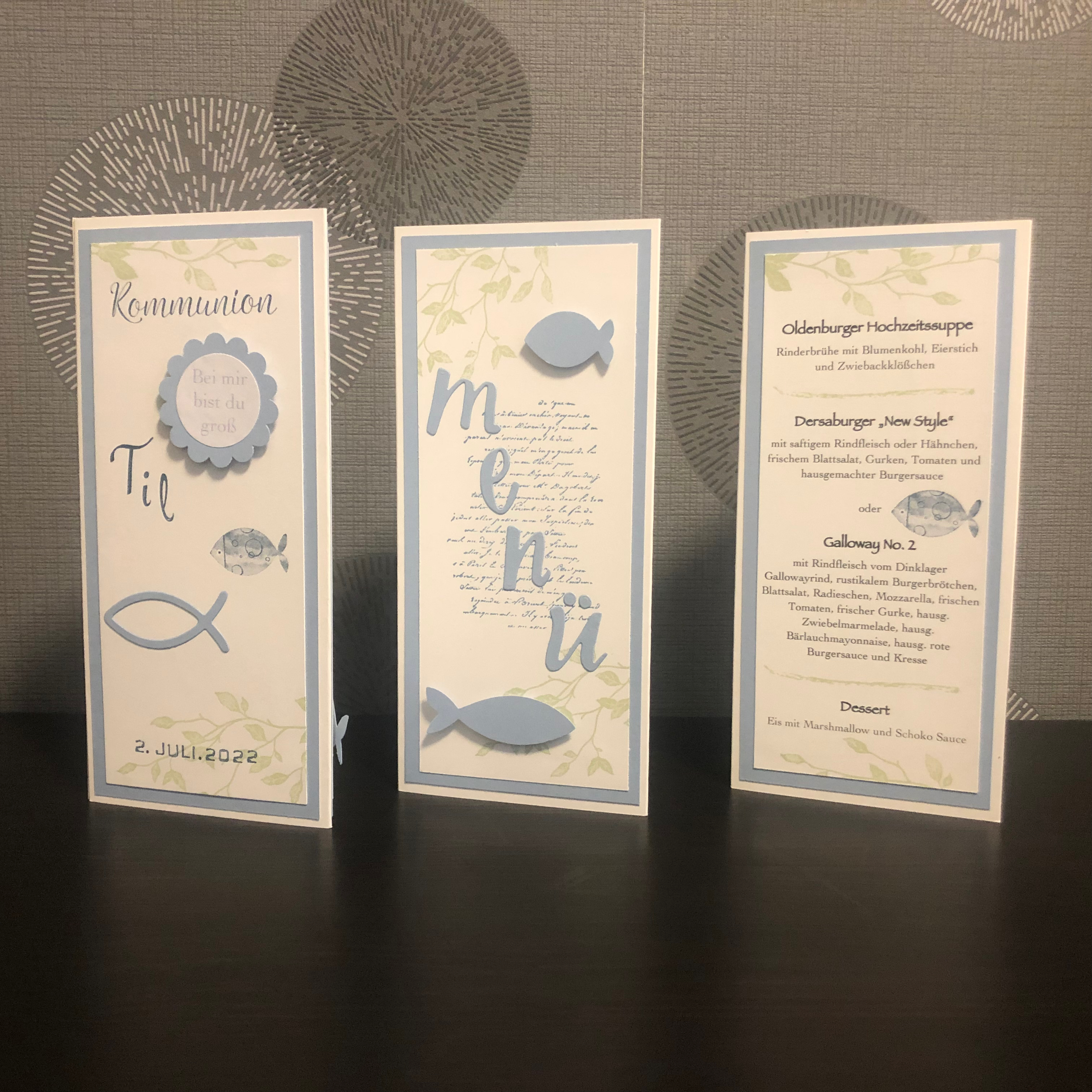 Zu sehen ist eine gebastelte Menükarte zur Kommunion in blau, weiß und grün für einen Jungen. Die Menükarte besteht aus drei Seiten. Die erste Seite ist beschriftet mit dem Namen des Kindes, das Datum der Kommunion und verziert mit zwei blauen Fischen. Die zweite Seite enthält das Wort Menü in ausgestanzten Buchstaben verzeiert mit zwei blauen Fischen und hinterlegt mit grünen Ranken und Schrift. Auf der dritten Seite ist das Menü gedruckt.