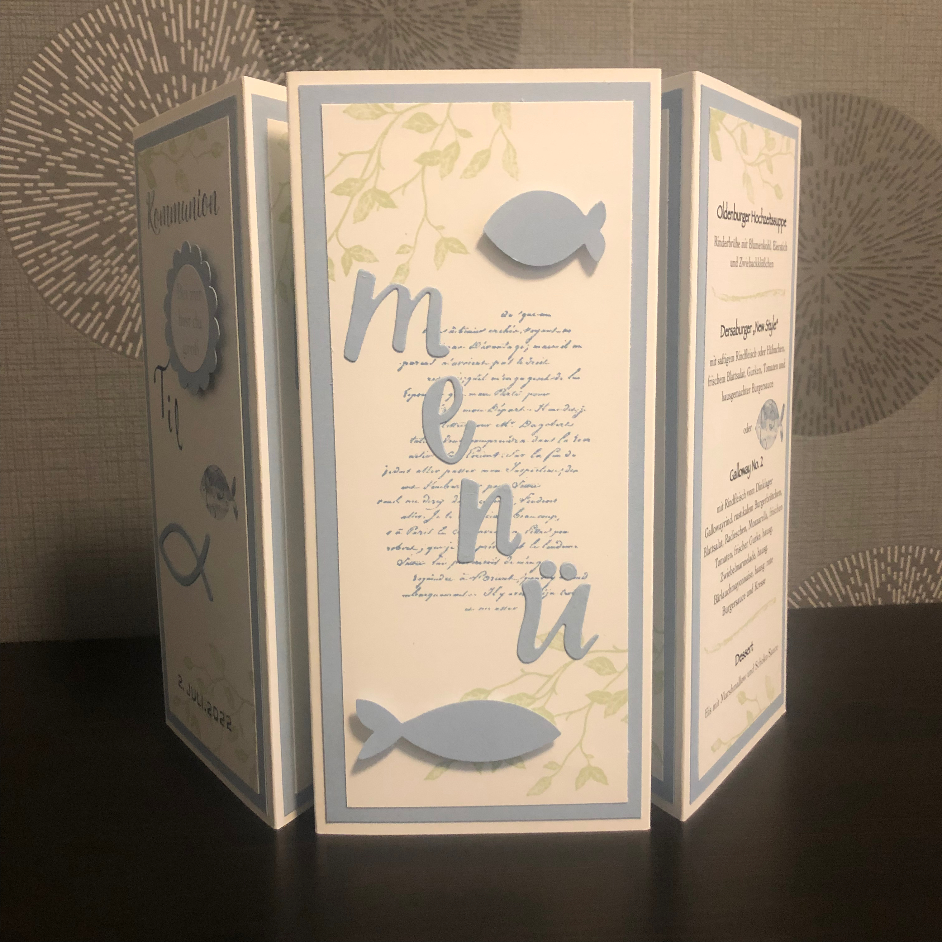 Zu sehen ist eine gebastelte Menükarte zur Kommunion in blau, weiß und grün für einen Jungen. Die Menükarte besteht aus drei Seiten. Die erste Seite ist beschriftet mit dem Namen des Kindes, das Datum der Kommunion und verziert mit zwei blauen Fischen. Die zweite Seite enthält das Wort Menü in ausgestanzten Buchstaben verzeiert mit zwei blauen Fischen und hinterlegt mit grünen Ranken und Schrift. Auf der dritten Seite ist das Menü gedruckt.