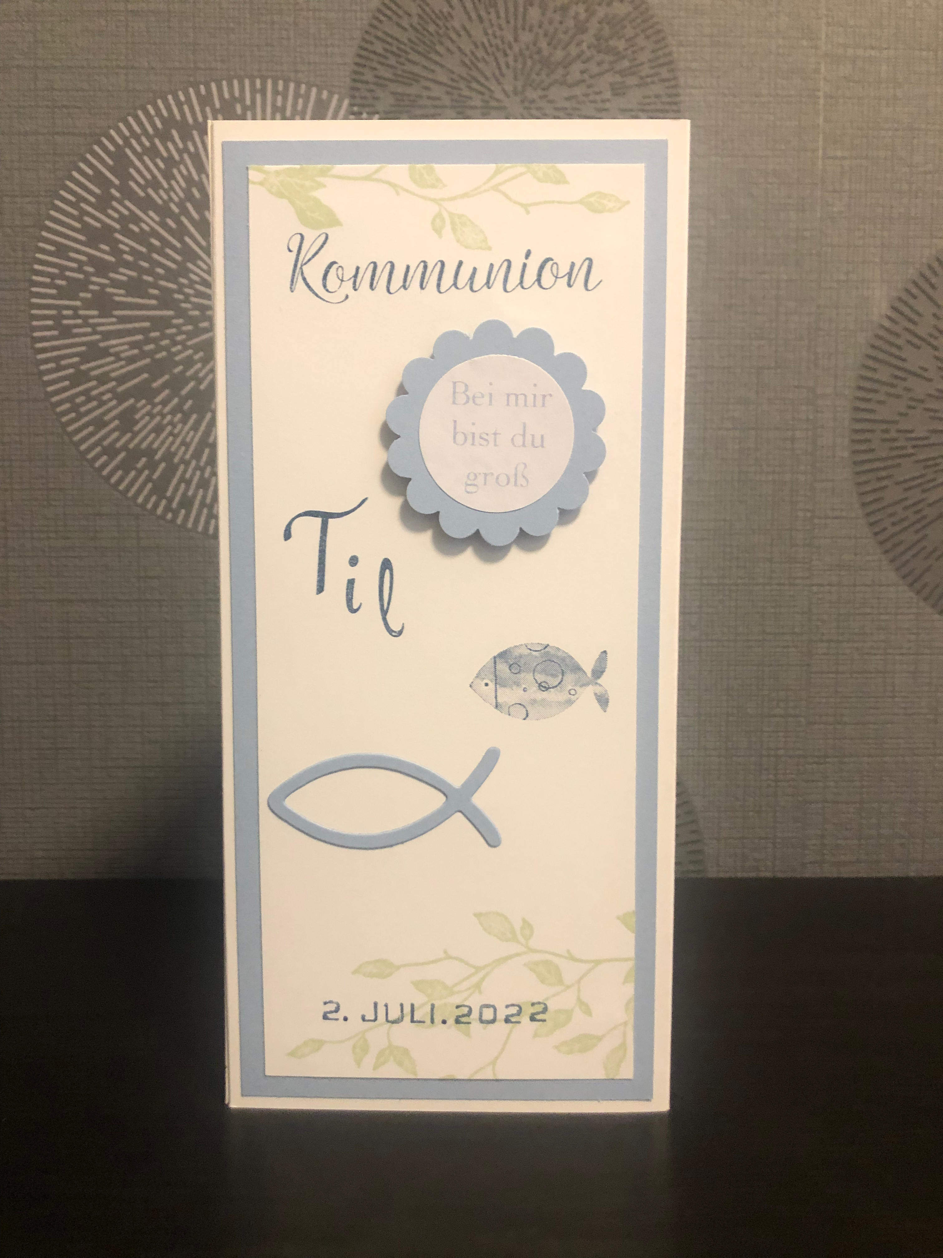 Zu sehen ist eine gebastelte Menükarte zur Kommunion in blau, weiß und grün für einen Jungen. Die Menükarte besteht aus drei Seiten. Die erste Seite ist beschriftet mit dem Namen des Kindes, das Datum der Kommunion und verziert mit zwei blauen Fischen. Die zweite Seite enthält das Wort Menü in ausgestanzten Buchstaben verzeiert mit zwei blauen Fischen und hinterlegt mit grünen Ranken und Schrift. Auf der dritten Seite ist das Menü gedruckt.