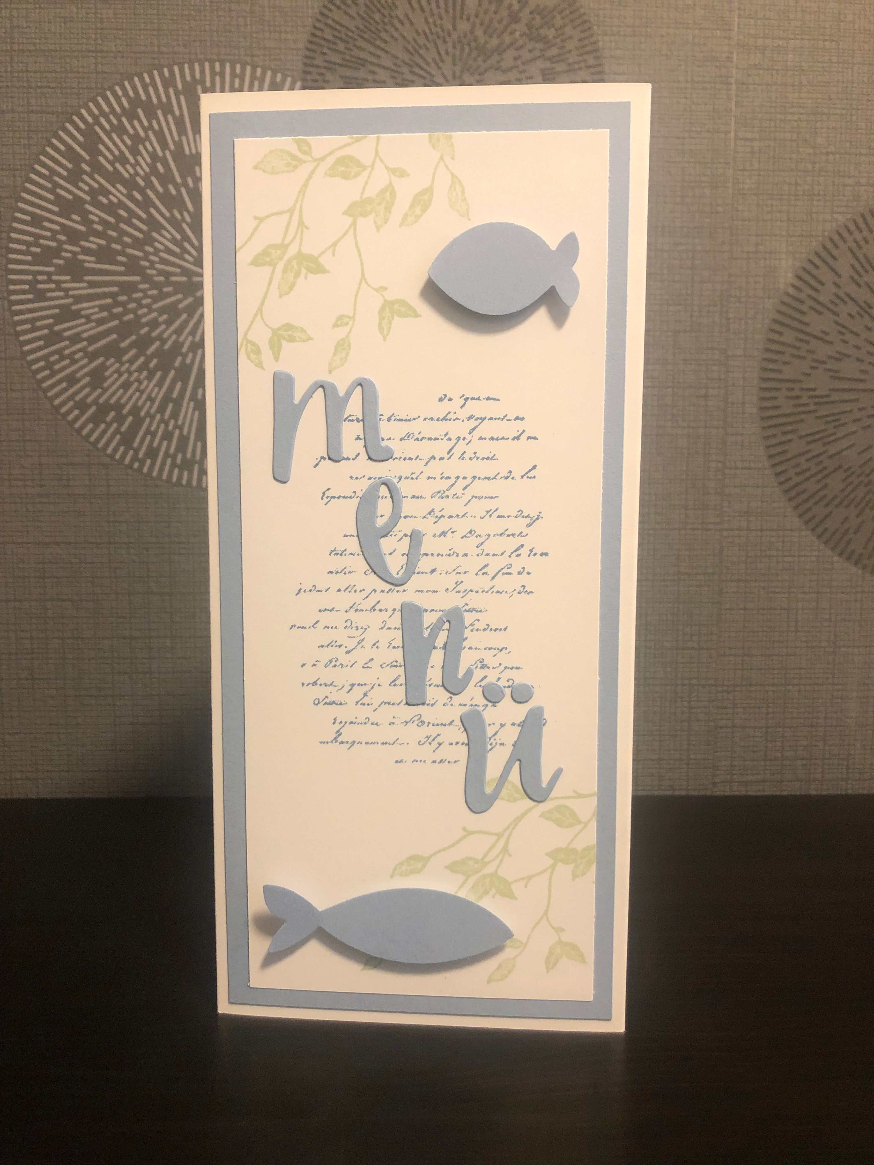 Zu sehen ist eine gebastelte Menükarte zur Kommunion in blau, weiß und grün für einen Jungen. Die Menükarte besteht aus drei Seiten. Die erste Seite ist beschriftet mit dem Namen des Kindes, das Datum der Kommunion und verziert mit zwei blauen Fischen. Die zweite Seite enthält das Wort Menü in ausgestanzten Buchstaben verzeiert mit zwei blauen Fischen und hinterlegt mit grünen Ranken und Schrift. Auf der dritten Seite ist das Menü gedruckt.