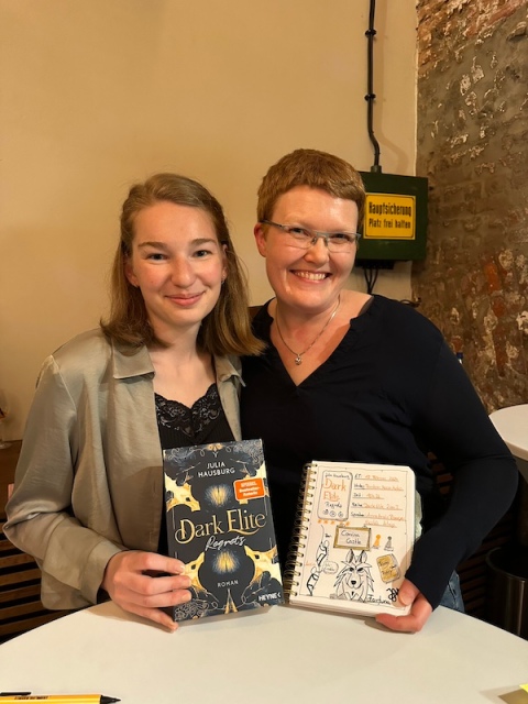 Ein Foto von Julia Hausburg und mir zusammen mit ihrem Roman Dark Elite Regrets und meinem Lesetagebuch