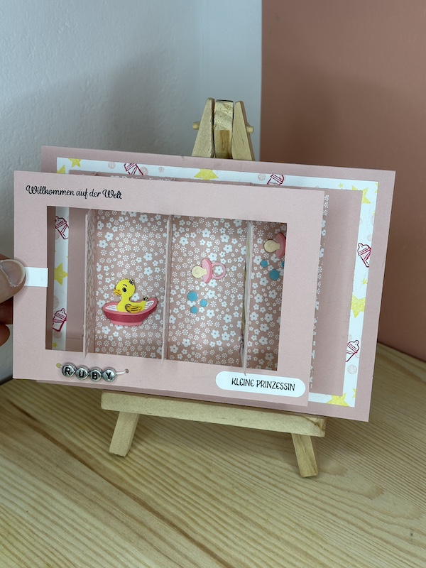 Babykarte in Rosa mit Slider für Mädchen. Vorne sieht man eine einen Elefanten und eine Giraffe jeweils auf Rollen. Darunter ist mit silbernen Kugeln der Name Ruby dargestellt. Zieht man an dem weißen Pfeil kann man dahinter Schnuller und Badeenten entdecken. Im Inneren befindet sich ein Glückwunsch zur Geburt