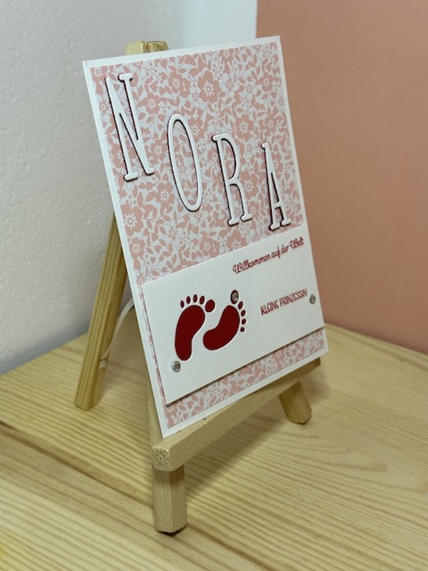 Zu sehen ist eine Babykarte mit dem Spruch: Willkommen auf der Welt kleine Prinzessin. Auf einem roas Hintergrund mit weißen Blümchen ist in Großbuchstaben der Name Nora zulesen. Darunter ist ein paar Babyfüsse ausgestanzt.