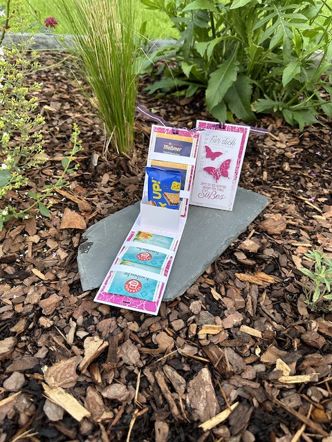 Auf dem Foto sieht man zwei kleine Verpackungen. Von der Größe passen vier Teebeutel und ein PickUp Schokokeks hinein. Außen sind Schmetterlinge aufgeklebt und oben ist die Verpackung mit einem Band verschlossen.