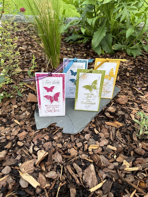 Auf dem Foto sieht man vier kleine Verpackungen. Von der Größe passen vier Teebeutel und ein PickUp Schokokeks hinein. Außen sind Schmetterlinge aufgeklebt und oben ist die Verpackung mit einem Band verschlossen.