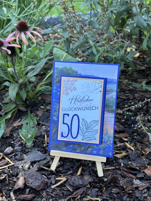 Zu sehen ist die Front einer Geburtstagskarte. Auf einem blauen Hintergrund mit Landschaftsmuster ist mittig ein weißes Rechteck geklebt auf dem eine schmale ausgestanzte 50 geklebt ist. Die Karte zum 50. Geburtstag in Blau ist zum Aufklappen.
