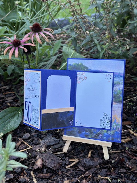 Zu sehen ist die Front einer Geburtstagskarte. Auf einem blauen Hintergrund mit Landschaftsmuster ist mittig ein weißes Rechteck geklebt auf dem eine schmale ausgestanzte 50 geklebt ist. An dem weißen Papier kann man ziehen und dann öffnet sich eine kleine Karte. Hier ist ein Steckfach für Geld und Gutscheinkarten zu sehen. Die Karte zum 50. Geburtstag in Blau ist zum Aufklappen.