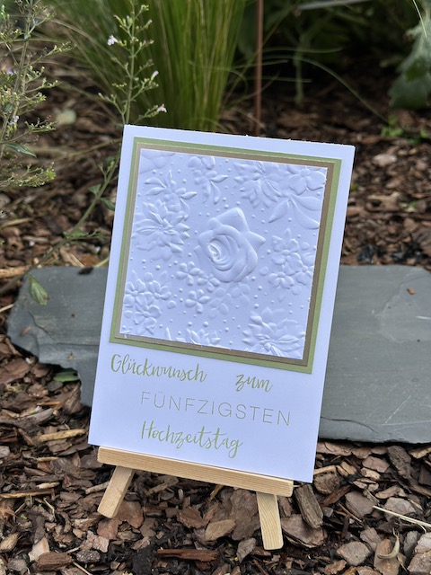 Zu sehen ist eine Karte zur goldenen Hochzeit. In weiß geprägt mit dem Prägefolder Feldblumen kann man eine Rose erkennen. Dahinter verbirgt sich eine goldene Mattung. Darunter steht: Glückwunsch zum fünfzigsten Hochzeitstag. Gebastelt mit Material von Stampin Up