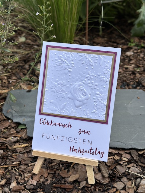 Zu sehen ist eine Karte zur goldenen Hochzeit. In weiß geprägt mit dem Prägefolder Feldblumen kann man eine Rose erkennen. Dahinter verbirgt sich eine goldene Mattung. Darunter steht: Glückwunsch zum fünfzigsten Hochzeitstag. Gebastelt mit Material von Stampin Up