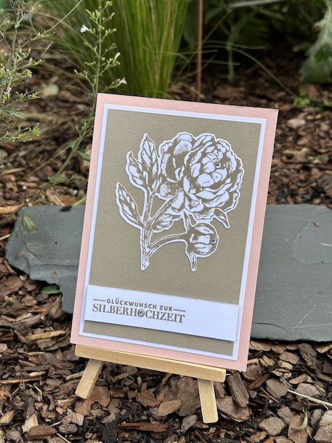 Zu sehen ist eine gebastelte Karte zur silbernen Hochzeit. Die Grundkarte ist rosa. Darauf ist eine Lage weißes und braunes Papier und darauf klebt eine in Savanne gestempelte und ausgestanzter Pfingstrose. Als Spruch steht unten auf der Karte: "Glückwunsch zur Silberhochzeit