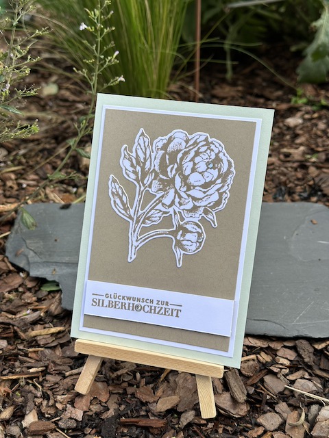 Zu sehen ist eine gebastelte Karte zur silbernen Hochzeit. Die Grundkarte ist grün. Darauf ist eine Lage weißes und braunes Papier und darauf klebt eine in Savanne gestempelte und ausgestanzter Pfingstrose. Als Spruch steht unten auf der Karte: "Glückwunsch zur Silberhochzeit