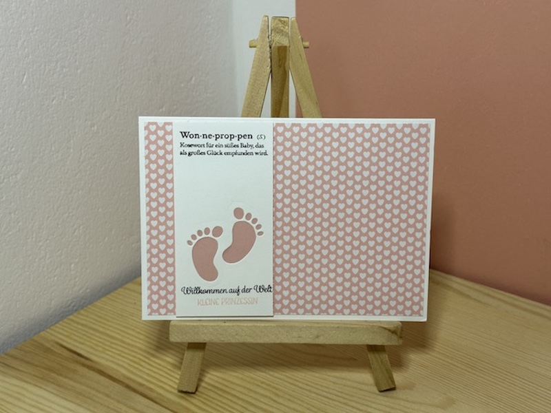 Zu sehen ist eine selbst gebastelte Babykarte in Rosa. Auf einem rosa Hintergrund ist ein weißer Streifen geklebt, aus dem ein Paar Babyfüße ausgestanzt ist, so dass man den rosa Hintergrund sehen kann. Als Spruch steht auf der Karte: Willkommen auf der Welt kleine Prinzessin.
