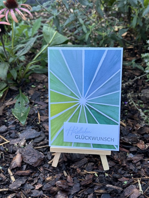 Zu sehen ist eine selbst gebastelte Karte. Der Hintergrund besteht aus weißen Strahlen, die sich vom MIttelpunkt in alle Richtungen ausbreiten. Die Dreiecke dazwischen verlaufen Farblich von gelb über grün nach blau. Darüber ist ein weißes Etikett geklebt, auf dem "herzlichen Glückwunsch" steht.