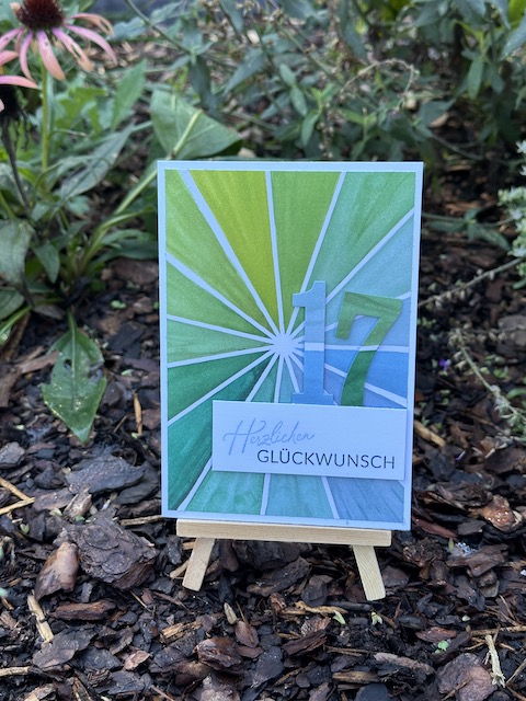 Zu sehen ist eine selbst gebastelte Karte. Der Hintergrund besteht aus weißen Strahlen, die sich vom MIttelpunkt in alle Richtungen ausbreiten. Die Dreiecke dazwischen verlaufen Farblich von gelb über grün nach blau. Darüber ist ein weißes Etikett geklebt, auf dem "herzlichen Glückwunsch" steht. Knapp hinter dem Etikett sind die Ziffern für 17 aufgeklebt, die Farblich an den Hintergrund angepasst sind.