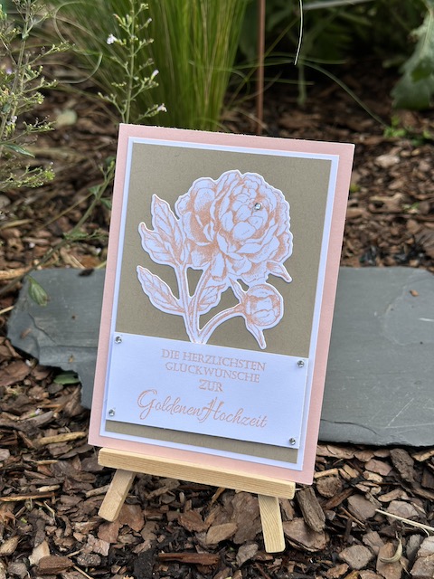 Zu sehen ist eine selbst gebastelte Karte zur goldenen Hochzeit. Eine rosa gestempelte Pfingstrose ist auf einen braunen Hintergrund, der sich durch einen weißen Rand von der Rosa Grundkarte abhebt. Zusätzlich ist auf der Karte noch folgender Text zu lesen: Die Herzlichsten Glückwünsche zur goldenen Hochzeit.
