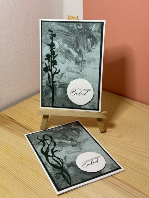 Zu sehen ist eine Trauerkarten dessen Hintergrund nach grünlichen Pinselstrichen aussieht. Davor ist ist in Schwarz ein ausgestanzter Streifen Blätter zu sehen. Daneben ist ein Wellenkreis angebracht mit dem Spruch: Stille Grüße von Herzen in dieser schweren Zeit