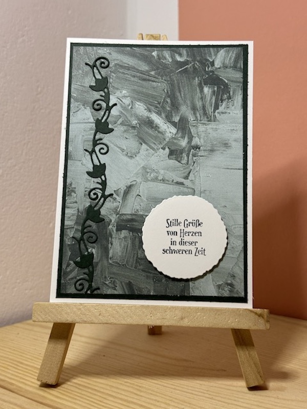 Zu sehen ist eine Trauerkarten dessen Hintergrund nach grünlichen Pinselstrichen aussieht. Davor ist ist in Schwarz ein ausgestanzter Streifen Blätter zu sehen. Daneben ist ein Wellenkreis angebracht mit dem Spruch: Stille Grüße von Herzen in dieser schweren Zeit