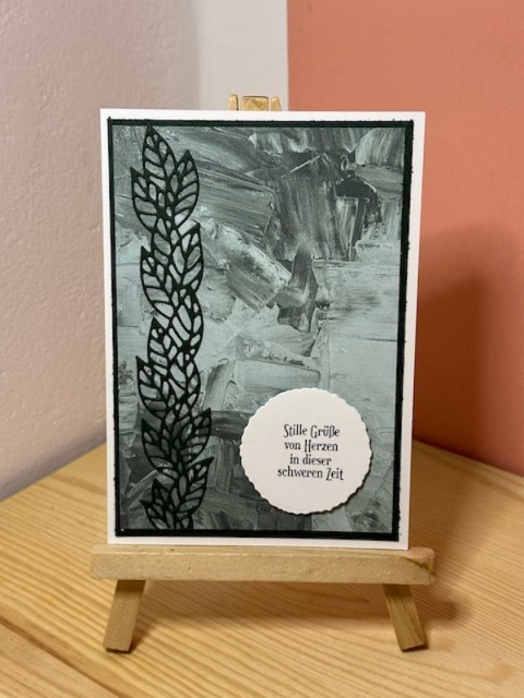 Zu sehen ist eine Trauerkarten dessen Hintergrund nach grünlichen Pinselstrichen aussieht. Davor ist ist in Schwarz ein ausgestanzter Streifen Blätter zu sehen. Daneben ist ein Wellenkreis angebracht mit dem Spruch: Stille Grüße von Herzen in dieser schweren Zeit