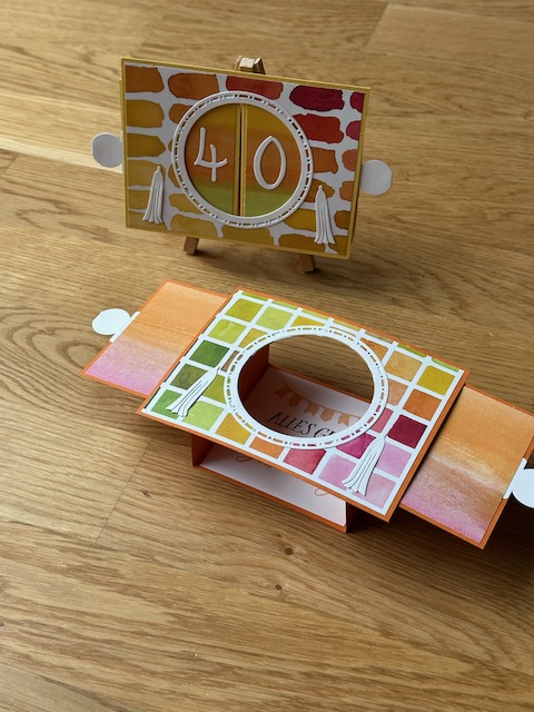Zu sehen ist eine Ziehkulissen Karte zum 40. bzw. 50. Geburtstag. Die Geburtstagskarte zeichnet sich dadurch aus, dass man an beiden Seiten ziehen kann und man dann durch den freien Kreis auf ein Bild dahinter Blicken kann. Ist die Karte auseinandergezogen, kann sie von alleine stehen. Farblich sind es gelb und rot Töne aus Material von Stampin Up gefertigt