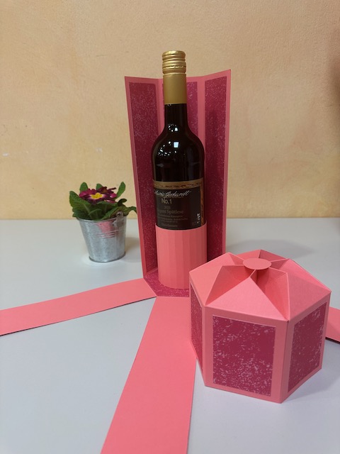 Zu sehen ist eine selbst gebastelte Verpackung für eine Weinflasche. Die Verpackung hat einen Deckel und drei Seitenteile klappen herunter, sobald man den Deckel öffnet. Es ist eine explosionsbox für eine Flasche. 