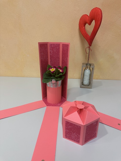 Zu sehen ist eine selbst gebastelte Verpackung für eine Weinflasche. Die Verpackung hat einen Deckel und drei Seitenteile klappen herunter, sobald man den Deckel öffnet. Es ist eine explosionsbox für eine Flasche.