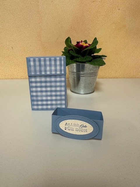 Zu sehen ist eine kleine selbstgebastelte Box in blau. Man kann die Banderole abziehen und dann die Box an der oberen Klappe aufmachen und durch aufrollen ganz öffnen. Im Inneren ist Platz für Genesungswünsche einen Teebeutel und Taschentücher und ein Hustenbonbon passt auch noch rein. 