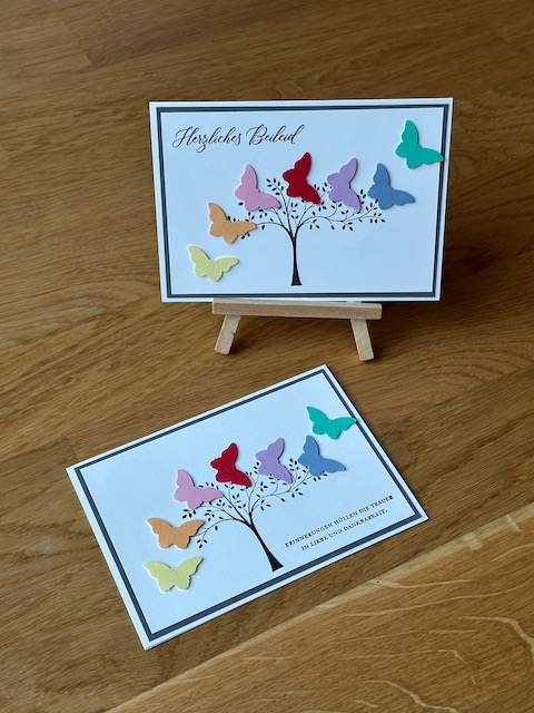 Zu sehen ist eine selbst gebastelte Trauerkarte. An einem gestempelten Baum fliegen scheinbar sieben bunte Schmetterlinge vorbei. Einzig ein schwarzer Rand hinter dem Bild greift das klassische Layout einer Trauerkarte auf.