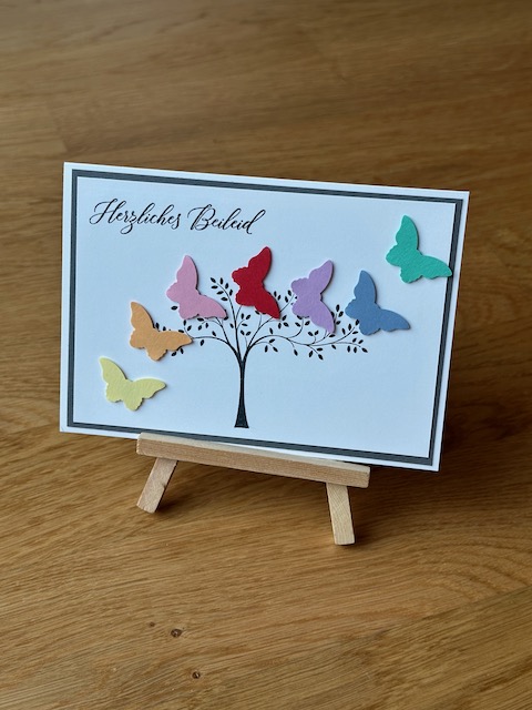Zu sehen ist eine selbst gebastelte Trauerkarte. An einem gestempelten Baum fliegen scheinbar sieben bunte Schmetterlinge vorbei. Einzig ein schwarzer Rand hinter dem Bild greift das klassische Layout einer Trauerkarte auf.
